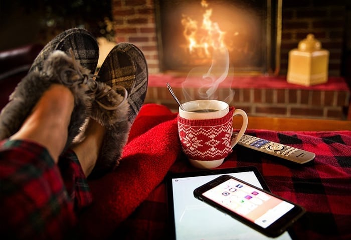 Person's feet kicked up on furniture next to a hot cup of liquid, a phone, and tablet in front of a fire place